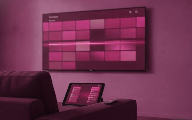 A television showing a channel guide grid, with a tablet and a phone listing channels on the sofa beside it