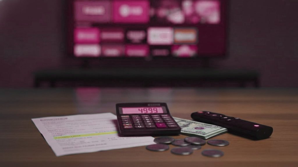 A calculator, dollar bills and coins next to a streaming stick and a remote on a table in front of a TV