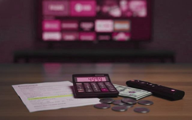A calculator, dollar bills and coins next to a streaming stick and a remote on a table in front of a TV